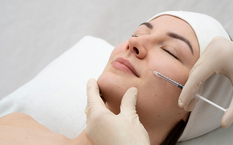 A imagem mostra uma mulher deitada em uma cama com um médico segurando uma seringa na frente do rosto. A mulher recebendo uma injeção facial de botox.
